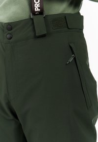 Donkergroene buitenbroek met ritszak, verstelbare klittenband tailleband en zwarte knoopsluiting op een lichtgroene fleece top.