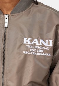 Brun bomberjakke med ribbet krave, lynlåslukning og hvidt broderet tekst, der lyder "KANI THE ORIGINAL EST. 1989 REG. TRADEMARK."