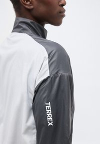 Προφίλ ατόμου που φοράει γκρι και λευκό μπουφάν με την επιγραφή "TERREX" στο μανίκι, βλέποντας προς τα δεξιά με απλό φόντο.