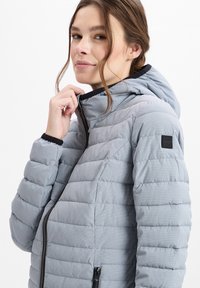 Padded jacke in hellgrau mit einem subtilen Karomuster. Verfügt über eine Kapuze, einen schwarzen Reißverschluss und Bündchen an den Handgelenken. Weicher Griff, leichtes Design.