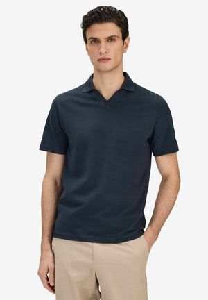 Man som bär mörkblå pikétröja med kort ärm och beige byxor, stående med ena handen i fickan mot en enfärgad vit bakgrund.