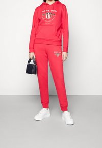 Sweat à capuche rouge et pantalon de survêtement en coton, avec une impression graphique et des logos. Assorti à des baskets blanches et un petit sac noir.