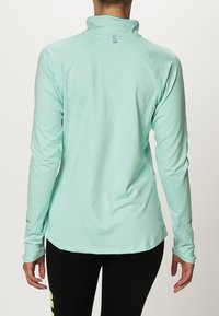 Kobieta ubrana w jasnoturkusowy, długi rękaw sportowego topu z otworami na kciuki, pokazana od tyłu, w połączeniu z czarnymi legginsami.