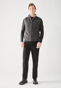 Gilet en tricot gris avec une fermeture éclair à l'avant, présentant un motif de câble texturé ; porté sur une chemise noire à manches longues et associé à un pantalon noir.