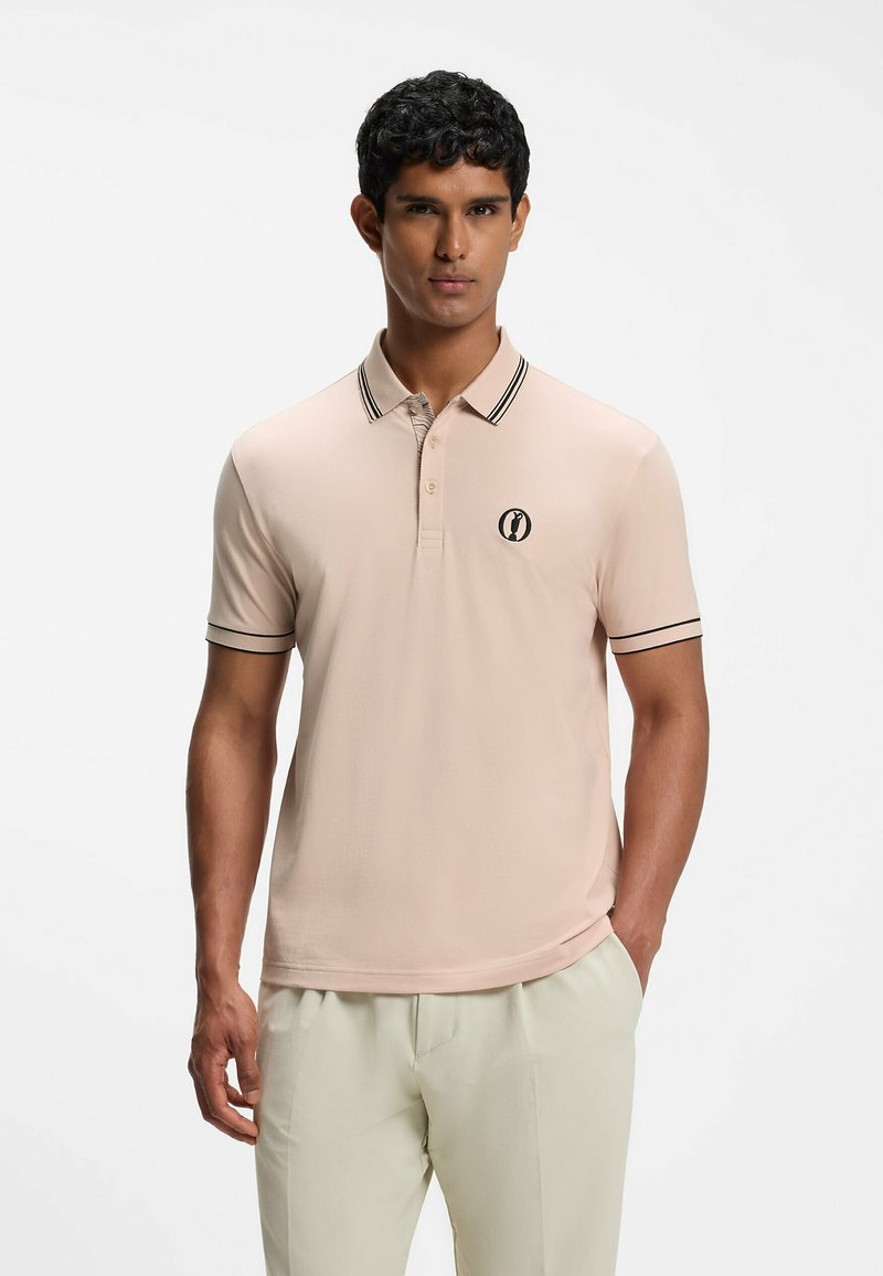Homme portant un polo rose pâle avec une bordure noire et un pantalon de couleur claire, debout devant un fond blanc uni.