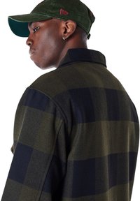 Groene geruite buitenkleding met zwarte vierkanten, gemaakt van textuurstof. Een groene corduroy pet completeert de look. Zilveren oorbel zichtbaar.