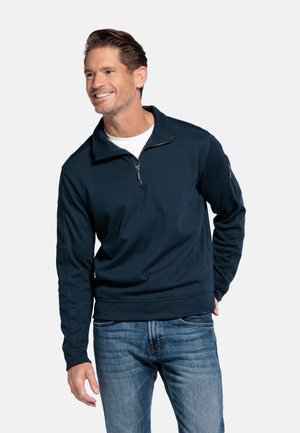 Marineblauer Pullover mit Reißverschlusskragen und strukturiertem Stoff. Ausgestattet mit Bündchenärmeln und einem geraden Saum, getragen über einem weißen T-Shirt.