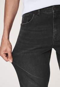 Czarne dżinsy z dżinsu o dopasowanym kroju i subtelnym efektcie sprania, z standardowym designem pięciu kieszeni i zapięciem na guziki z przodu, trzymane jedną ręką.