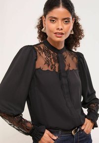 Must värviline pluus, millel on õhuke pitsdetail rindade ja varrukate piirkonnas, volangäär ja paksu varrukad. Pehme materjal, millel on figuuri rõhutav siluett.