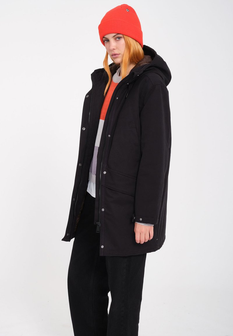 Giacca nera lunga con cappuccio, zip frontale e bottoni a pressione. Abbinata a un maglione a righe colorato e pantaloni neri. Berretto arancione brillante in testa.