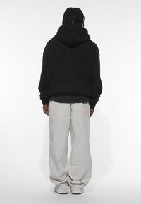 Fekete kapucnis pulóver, laza fazonban, bordázott mandzsettákkal és szegéllyel, világosszürke bő nadrággal párosítva, oldalzsebekkel és rugalmas nadrágszárakkal.