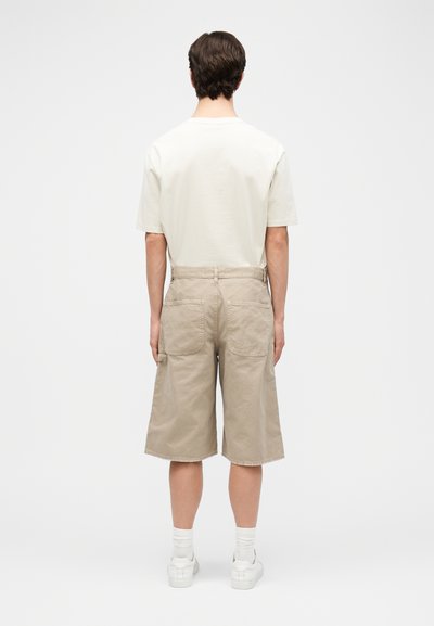 Homme portant une chemise blanche à manches courtes, un short beige mi-genou avec des poches arrière, des chaussettes blanches et des baskets blanches, debout de dos.
