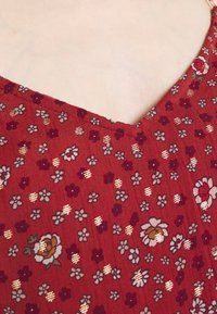 Tessuto rosso con motivi floreali, caratterizzato da piccoli fiori in tonalità di bianco, giallo e viola, accentuato da strisce dorate. Design a V.