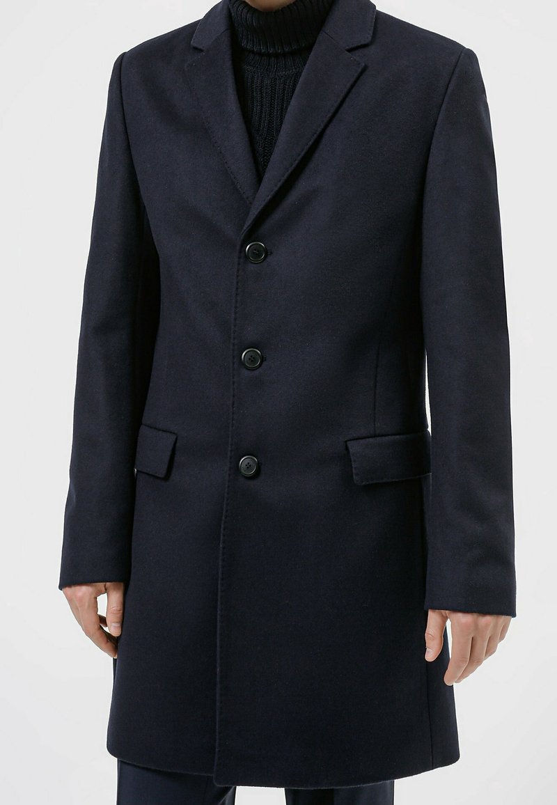 Navy uldblandet overfrakke med en hakrevers, lukning med fem knapper, sidelommer og lige snit. Tekstureret stof giver varme.
