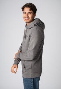 Grijze hoodie met trekkoord, logo op de capuchon, geribbelde manchetten en zak. Draagt men met blauwe jeans, wat een casual, aansluitende uitstraling geeft.