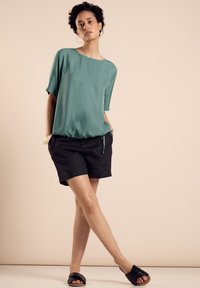 Teal blouse with short sleeves, gathered hem, paired with black shorts. Model wears black slide sandals and simple earrings. Neutral background.