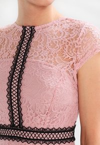 Rosa spetsklänning med svarta dekorativa kanter och detaljer. Korta ärmar med blommönster och texturerad finish runt halsringningen.