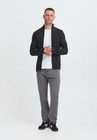 Jonge man staat in een zwarte ritstrui, witte T-shirt, grijze jeans en zwarte sneakers met witte strepen tegen een effen achtergrond.