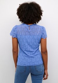 Haut en dentelle bleue avec un motif floral, manches courtes, encolure ronde, et une ouverture en forme de goutte dans le dos, associé à un jean en denim.