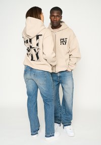 Sudaderas con cremallera beige y gran texto negro en la parte posterior, combinadas con jeans azul claro y zapatillas blancas. Vestimenta casual con un ajuste holgado.