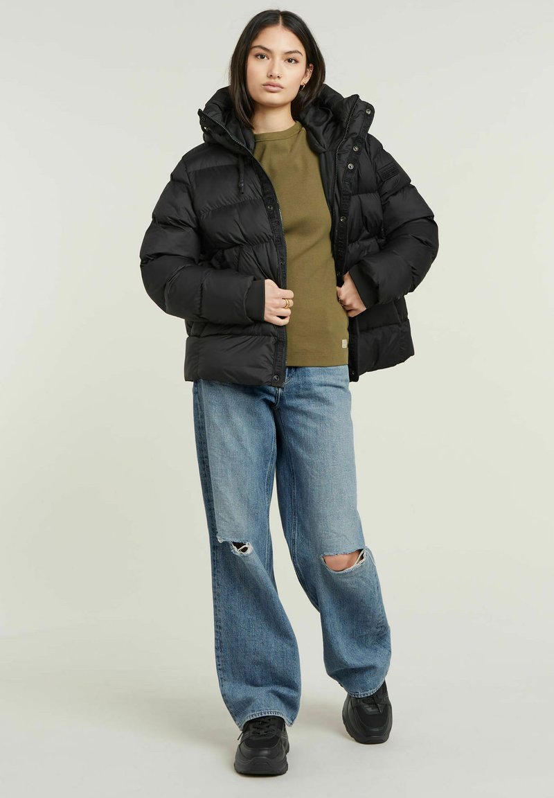 Schwarze gepolsterte Jacke mit Kapuze, ausgestattet mit einem gesteppten Design. Über einem olivgrünen Oberteil getragen und kombiniert mit hellen, ausgewaschenen Jeans und schwarzen Schuhen.