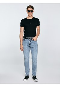 Noor noormees, kes kannab musta päikeseprille, musta lühikeste varrukatega särki, helesiniseid teksapükse, musta vööd ja musti tossusid, seisab ühevärvilise tausta ees.