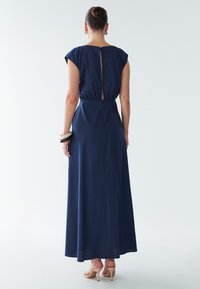 Tamsiai mėlyna, be rankovių maxi suknelė su atvira nugaros dalimi ir elastinga juosmens linija. Sklandi medžiaga ir laisva silueto linija. Dėvima su smėlio spalvos sandalais.