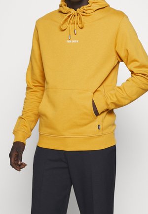 Personne portant un sweat à capuche jaune moutarde avec le texte « LES DEUX » sur la poitrine, main dans la poche avant, associée à un pantalon noir.