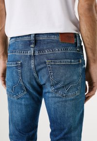 Mörkblå denimjeans med en läderlogotyp, två bakfickor med kontrasterande sömnad och ett unikt V-format mönster på klaffarna.