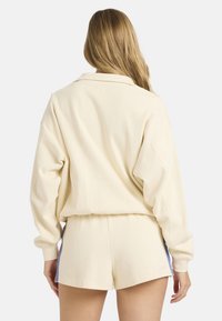 Φαρδιά φούτερ σε κρεμ απόχρωση, κατασκευασμένο από μαλακό ύφασμα, με γιακά και χαλαρούς ώμους, συνδυασμένο με σορτς στο ίδιο χρώμα.