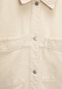 Giacca color crema con chiusura frontale a bottoni, due grandi tasche sul petto, bottoni argento e tessuto strutturato e resistente.