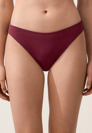 Ženski trup z teksturiranimi temno rdečimi spodnjicami bikinija, ki prikazujejo srednji del telesa in zgornji del stegen na nevtralnem ozadju.