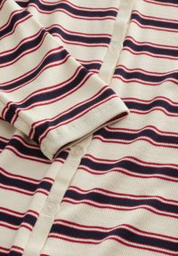 Cardigan côtelé crème avec des rayures horizontales bleu marine et rouge. Fermeture à cinq boutons avec une texture douce et des poignets repliés.