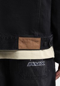 Zwarte denim jas met een bruine leren patch met reliëftekst. De stof toont een subtiele textuur. Daaronder is een crèmekleurig shirt zichtbaar.