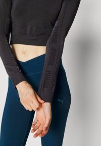 Svart crop top med lång ärm som har upphöjd "PUMA"-text på ärmarna; kombinerad med mörkblå leggings och subtil logotypdetalj.