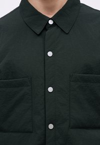 Camisa de botão verde-escura com gola, botões de pressão prateados e dois bolsos frontais com uma textura enrugada.