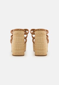 Un par de sandalias de cuña color canela con tacones trenzados y dobles tiras en los tobillos, vistas desde la parte posterior sobre un fondo blanco.