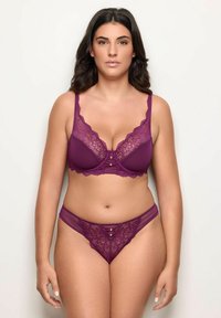 Femme à la peau mate et aux cheveux foncés posant en lingerie assortie en dentelle violette, composée d'un soutien-gorge et d'un slip, sur un fond clair uni.