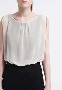 Femme portant un haut sans manches en chiffon blanc avec des plis au niveau du col et une jupe noire texturée devant un fond blanc.