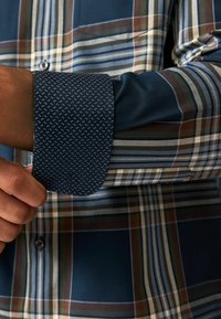 Camisa de vestir a cuadros en azules, verdes y marrones, con un puño y detalle de botones de lunares en azul marino que contrastan. Textura de tela suave.