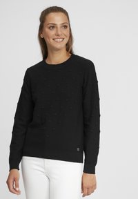 Pulover negru cu o suprafață texturată, având puncte ridicate. Decupaj rotund la gât și mâneci lungi. Asortat cu pantaloni albi. Design simplu.