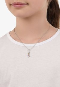 Silverhalsband med en hängande berlock som har färgglada ädelstenar, bärs över en vit T-shirt med rundad halsringning.