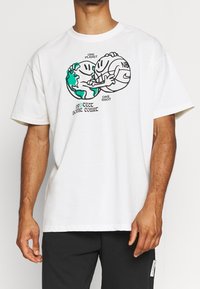 Bílá bavlněná tričko s krátkým rukávem, na kterém je grafický design dvou kreslených postaviček objímajících globus a basketbalový míč. Text zní "JEDNA PLANETA JEDNA ŠANCE."