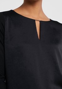 Sort top med rund hals og en nøglerækkeudskæring, sikret med en lille metallisk accent. Glat tekstur med lange ærmer.