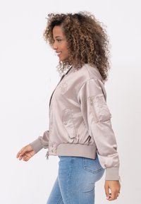 Lichtroze satijnen bomberjack met ribbelzoom, ritszakken en geborduurde details op de borst; gecombineerd met blauwe jeans.