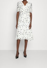 Robe blanche avec un design croisé, des manches courtes bouffantes et une ceinture à la taille. Décorée de pois bleus et gris.