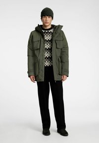 Man med grön mössa, olivgrön huva parka, svartvit zigzagtröja, svarta byxor och svarta skor som står på vit bakgrund.