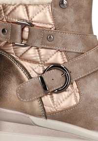 Zapatilla cuña beige con panel de satén acolchado, dos correas ajustables con hebillas plateadas y un detalle de botón metálico.