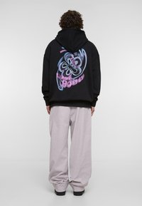 Musta huppari, jossa värikäs graafinen kuvio selässä, abstrakti kukkakuosi sinisen ja vaaleanpunaisen sävyissä. Yhdistetty löysiin vaaleanharmaisiin housuihin.