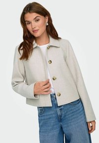 Donna che indossa una giacca beige chiaro con bottoni sopra una maglietta bianca e jeans blu, in posa con la mano vicino all'apertura della giacca su sfondo bianco.